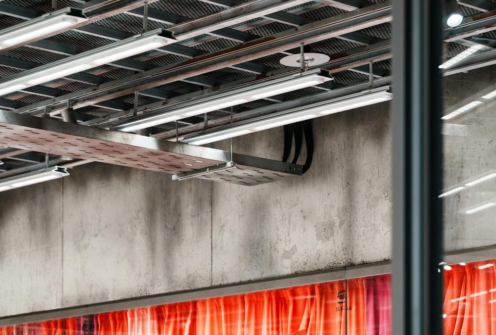 Exposed commercial ceiling with metal cable trays, light fixtures and conduit along a concrete wall