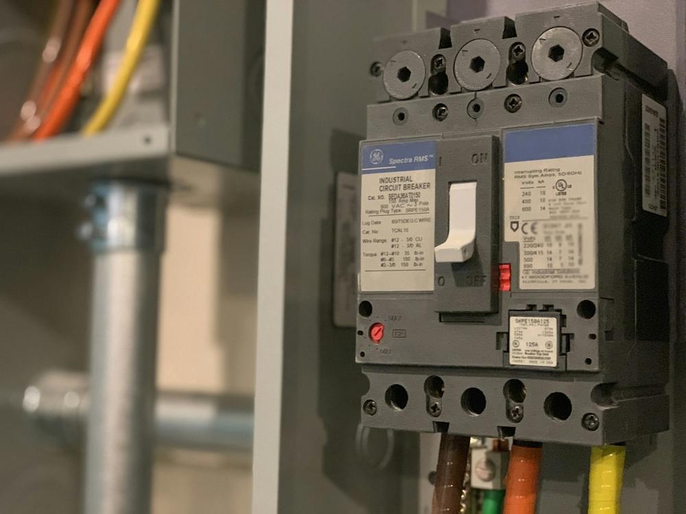 Gray industrial circuit breaker mounted in a panel with metal conduit and color-coded wiring
